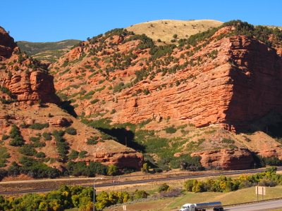 [Red segmented rock has green vegetation growing in rows across it where there are ledges. The very top of the rock is grass-covered. All this dwarfs the roadway at the base of the walls.]